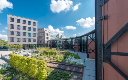 Projekte_Slider_Gallery_Lokschuppen_09 Lokschuppen mit Glasfassade neben Neubau und Park mit Brunnen, Büschen, Schienen